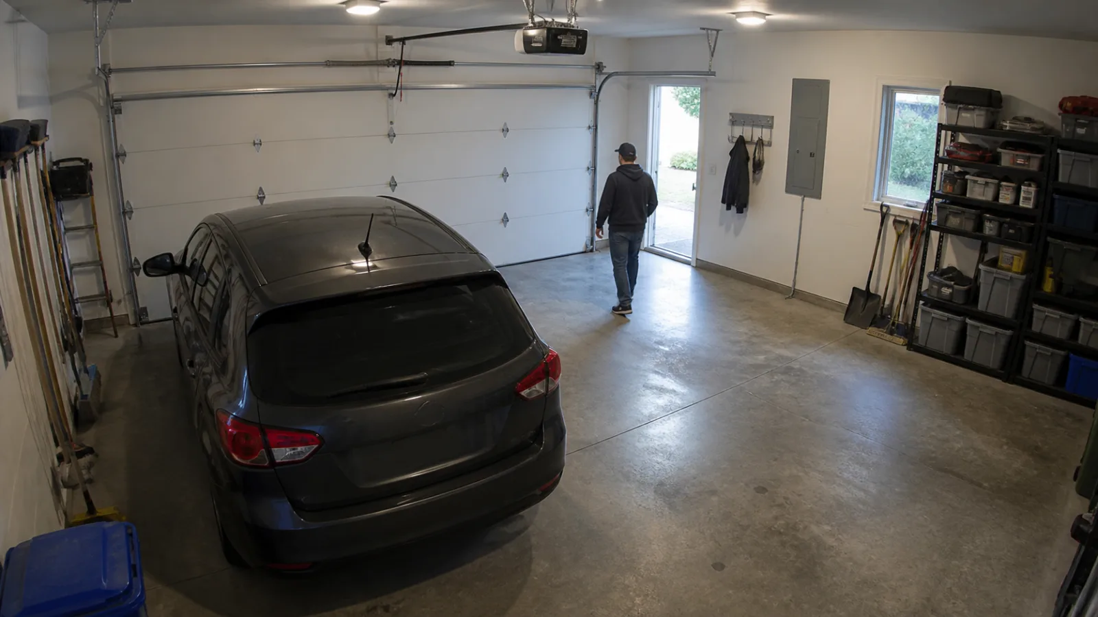 Forhøjet fast sikkerhedskameraudsigt inde i en boliggarage med en hoveddør, sideindgang, parkeret bil, hylder og en fjern person